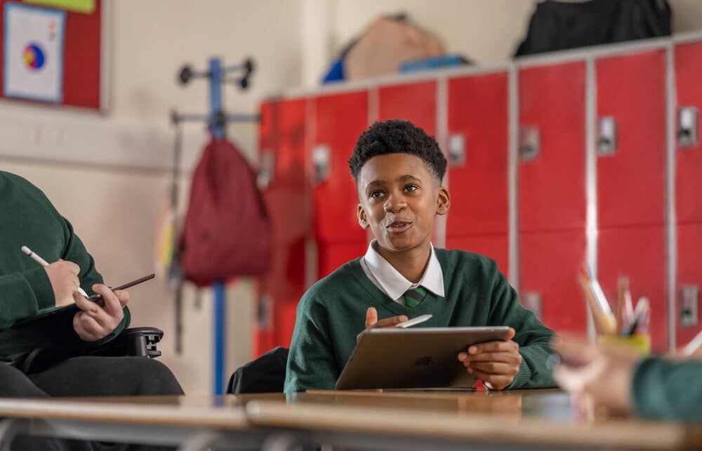 Student smiling whilst using an iPad with an Apple Pencil in a classroom.