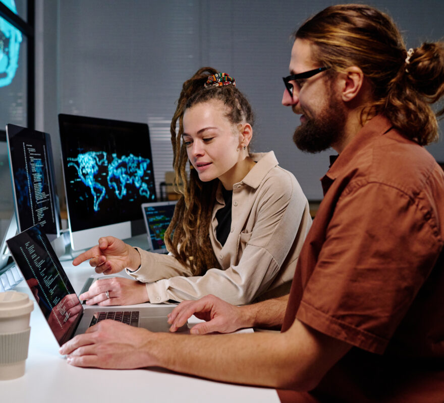 Young confident female programmer pointing at laptop screen with coded data while exlaining something to male colleague at night meeting